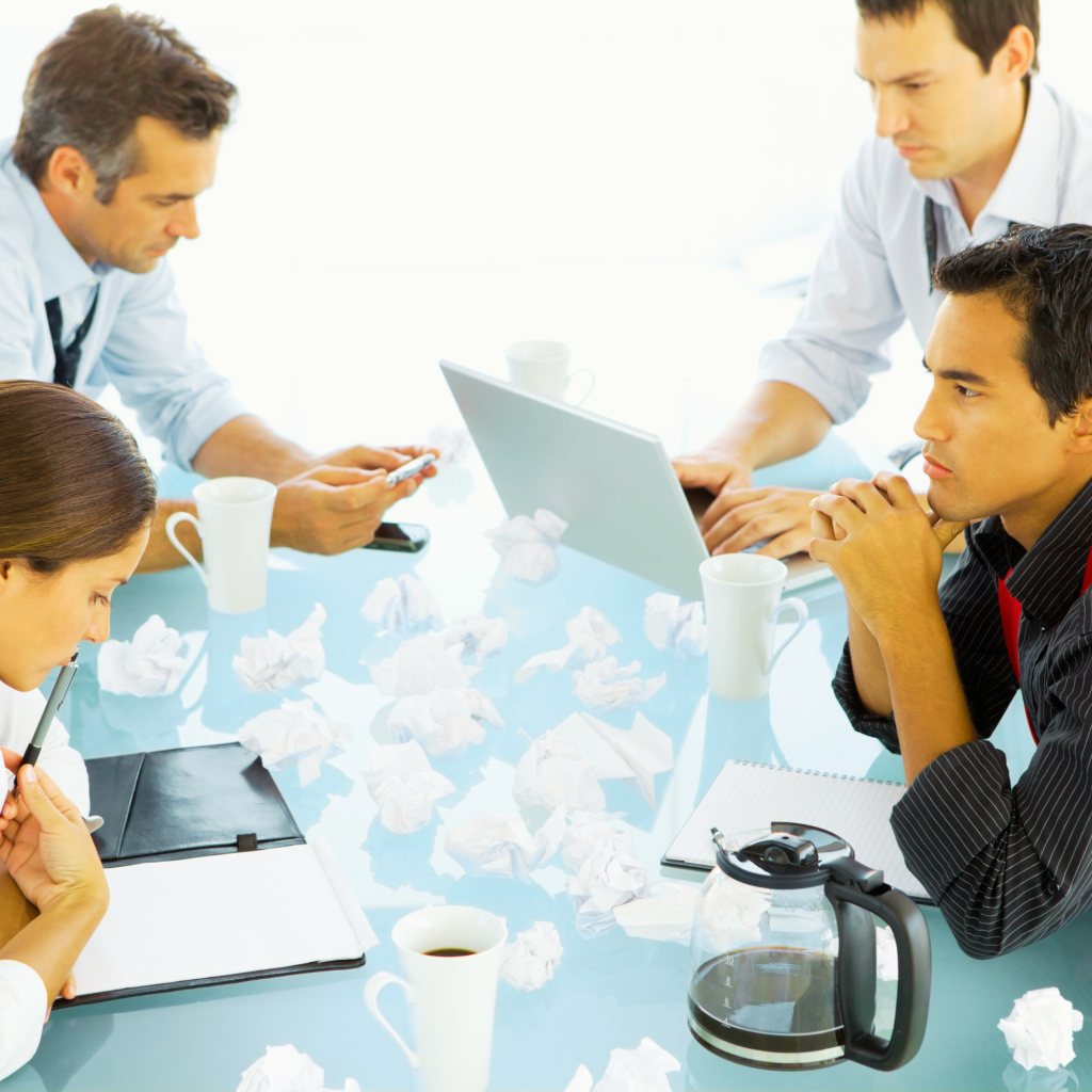 A group of professionals sitting around a table, brainstorming with laptops, notebooks, and scattered crumpled paper.